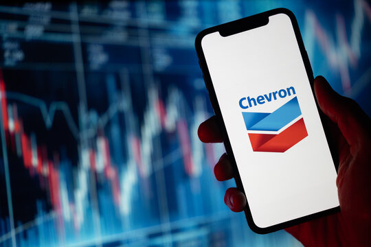 In silhouette, a hand holds a mobile phone with the Chevron logo displayed. Behind it, a backdrop illustrates descending stock prices highlighted in red color