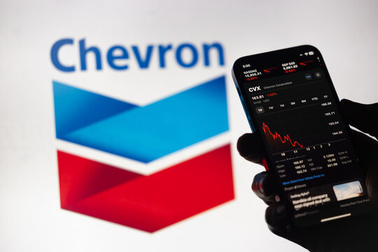 A backlit hand holds a phone, showcasing red descending Chevron stock prices within the Apple stock market app. In the backdrop, a blurred screen of the logo