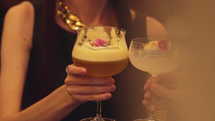 Closeup cropped woman hand making a toast mixed passion fruit smoothie cocktail wearing ring with friend at blurred drink background at nighttime warm light at nightclub counter bar party. Vinosity.