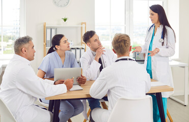 Group of professional doctors sitting with laptop at the desk on meeting discussing diagnosis, checking test results or planning treatment in clinic. Medical staff, healthcare and teamwork concept.