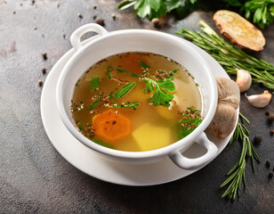 A white bowl of clear broth soup with sliced carrots and fresh parsley is surrounded by celery stalks, garlic cloves, and spices on a gray stone surface.