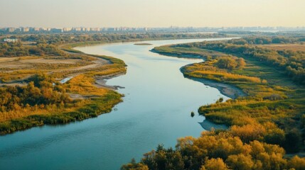 Obraz premium Serene View of a Meandering River Surrounded by Lush Green Vegetation under a Clear Sky with Urban Skyline in the Distance at Sunset