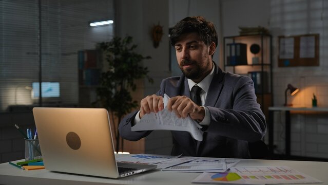 Adult business man manager sitting at desk working on laptop in office in evening, meeting online by video call, ripping papers with infographic.