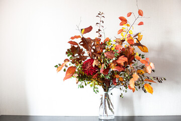 Multicoloured autumn bouquet on a table. Bunch of colorful fall branches and red chrysanthemum flower in glass vase at home interior. Floral composition, make DIY bouquet.