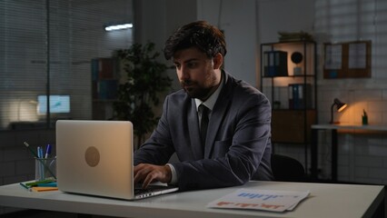 Adult business man manager sitting at desk working on laptop in office in evening, typing on computer doing research, reading information.