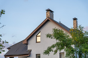 A modern house showcases a distinctive roof and chimney, surrounded by greenery under a clear sky during the evening hours, highlighting its architecture