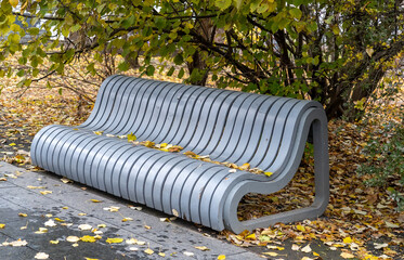 A sleek gray bench is covered in fallen leaves, set against a backdrop of green and yellow foliage in a peaceful park during autumn