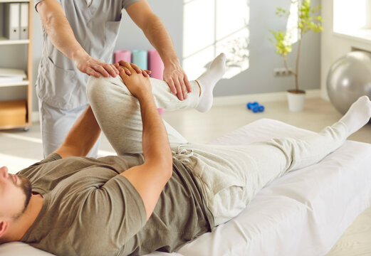 Male physician or osteopath massaging and stretching patient leg during rehabilitation in clinic. Physiotherapist doing manual therapy and healing treatment for knee recovery to young man on couch.