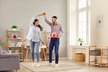 Fototapeta premium Happy loving couple at home, having fun in living room, dancing together, celebrating new flat or anniversary, handsome young husband holding beautiful wife hand, moving enjoying date, gift box giving