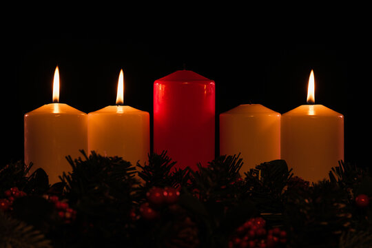 four white advent candles in a row where three are lit, with a red unlit Christ candle in the center, and garland below. 