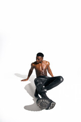 A young African American man sits confidently on the floor, bare chested, wearing stylish black pants.