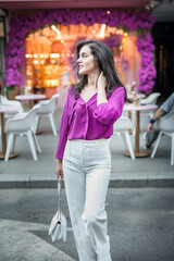 Young beautiful brunette woman in a pink jacket and white pants posing on the street. Portrait of a beautiful young woman in a purple blouse and white pants posing in an outdoor cafe on the street