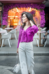 Young beautiful brunette woman in a pink jacket and white pants posing on the street. Portrait of a beautiful young woman in a purple blouse and white pants posing in an outdoor cafe on the street