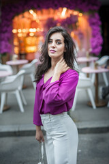 Fototapeta premium Young beautiful brunette woman in a pink jacket and white pants posing on the street. Portrait of a beautiful young woman in a purple blouse and white pants posing in an outdoor cafe on the street