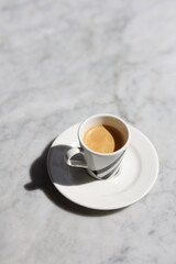 A freshly brewed espresso served in a white porcelain cup on a matching saucer, placed on a marble table with natural lighting.