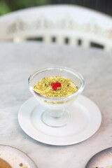 Traditional Middle Eastern dessert topped with crushed pistachios and garnished with a bright red cherry, elegantly served in a glass dish on a marble table.
