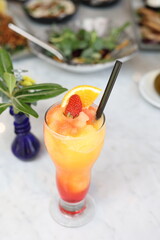 Vibrant layered fruit smoothie garnished with fresh strawberry and orange slice, served in a tall glass on a marble table with an assortment of dishes in the background.