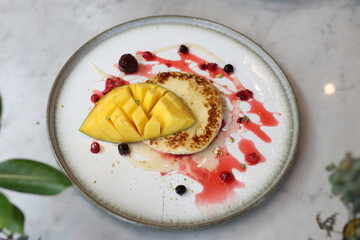 Delicious pancake topped with fresh mango slices, drizzled with berry sauce and honey, garnished with vibrant berries on a white ceramic plate.
