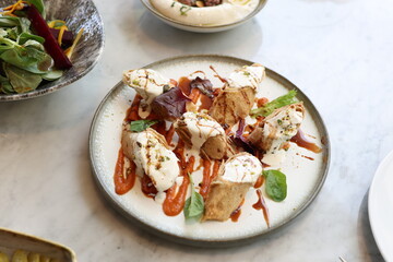 A gourmet Middle Eastern dish featuring crispy stuffed rolls topped with creamy yogurt and pistachios, drizzled with roasted pepper sauce and garnished with fresh greens.