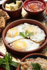 A rustic Lebanese breakfast featuring sunny-side-up eggs garnished with mint leaves, served alongside zaatar flatbread, butter, apricot jam, and chickpea hummus, presented in ceramic bowls.