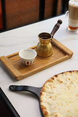 Traditional Turkish coffee served in a brass cezve with a ceramic cup on a wooden tray, alongside a cheesy bread and a latte in the background.