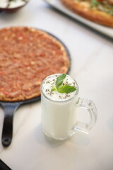 A traditional Lebanese manoushe topped with seasoned minced meat, served on a black tray beside a glass of laban yogurt drink, presented on a marble table.