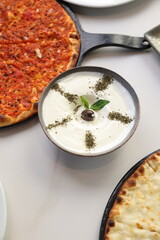A traditional Lebanese breakfast spread featuring tomato manoushe, cheese manoushe, and a bowl of creamy labneh garnished with an olive and dried herbs, served on a rustic setup.