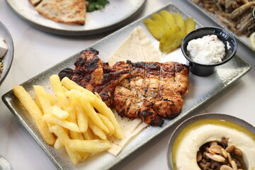 Close-up of a grilled chicken breast, perfectly charred and juicy, served alongside crispy golden French fries. The dish is accompanied by a small black bowl of creamy garlic sauce.