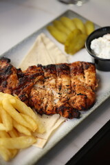 Close-up of a grilled chicken breast, perfectly charred and juicy, served alongside crispy golden French fries. The dish is accompanied by a small black bowl of creamy garlic sauce.