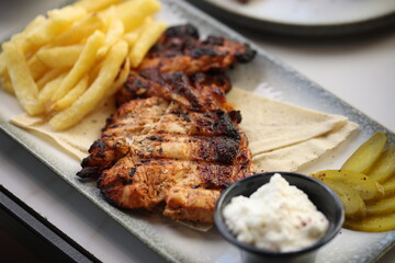 Close-up of a grilled chicken breast, perfectly charred and juicy, served alongside crispy golden French fries. The dish is accompanied by a small black bowl of creamy garlic sauce.