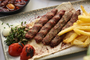 A platter of chargrilled Lebanese beef kebabs served with crispy French fries, grilled tomatoes, onions, fresh parsley, and sumac seasoning on flatbread, offering a hearty and flavorful Middle Eastern