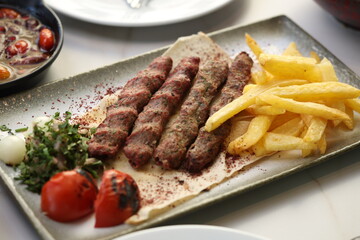 A platter of chargrilled Lebanese beef kebabs served with crispy French fries, grilled tomatoes, onions, fresh parsley, and sumac seasoning on flatbread, offering a hearty and flavorful Middle Eastern