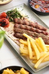A platter of chargrilled Lebanese beef kebabs served with crispy French fries, grilled tomatoes, onions, fresh parsley, and sumac seasoning on flatbread, offering a hearty and flavorful Middle Eastern