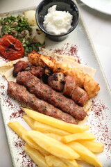 A Lebanese mixed grill platter featuring chargrilled beef kebabs, chicken skewers, and lamb skewers, served with French fries, garlic sauce, grilled tomatoes, fresh parsley, and sumac on flatbread, of