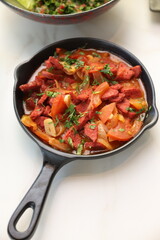 A skillet of spicy Lebanese soujouk sausages cooked with sautéed onions, tomatoes, and garlic in a rich, tangy tomato-based sauce, garnished with fresh parsley, delivering a bold and flavorful Middle 