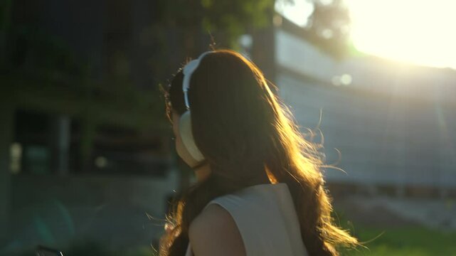 Young carefree asian woman in casual style using smartphone with headphone enjoy listening music and walking along the public park through morning sunlight, happy female in urban lifestyle concept.