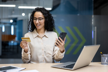 Hispanic business woman in office holding smartphone and credit card, demonstrating modern technology use for online shopping. Laptop on desk indicates digital work environment.