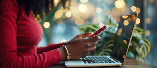 A young black woman is sitting in a cafe, using her laptop and smartphone. She is wearing a red sweater and has her hair in a ponytail.
