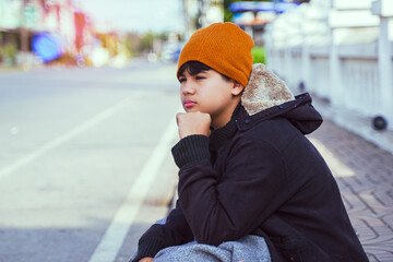 A teenage boy wearing a black jacket and yellow beanie sits on the sidewalk. Put your hands on your...