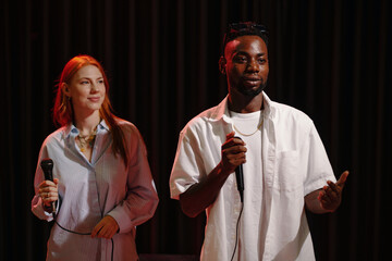 Portrait of African American man and Caucasian woman presenting on stage holding microphones...