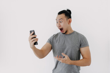 Shock and wow face Asian man in grey t-shirt use smartphone app on white.