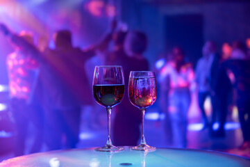 Colorful nightclub atmosphere with focus on two wine glasses while people dance in background. Perfect for nightlife themes.