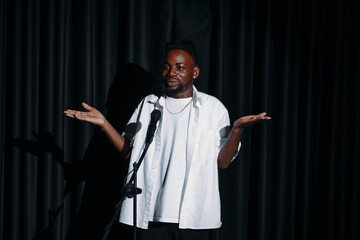Male comedian standing on stage with microphone, smiling and performing under spotlight. Wearing casual white shirt, engaging with audience while raising hands