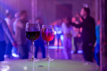 Two wine glasses on a table at a lively party with colorful lights and people dancing in the background.
