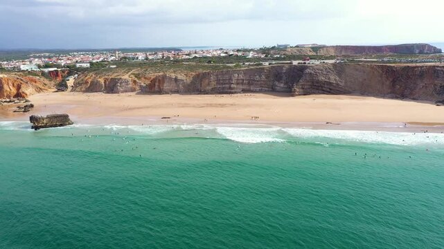 Hundreds of surfers in the water, in South of Portugal, Sagres, Tonel Beach. Aerial drone view. Turquoise water. Famous travel destination. High cliff. Drone forward, descending and camera tilt up.