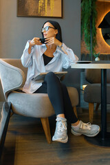 A woman with dark hair and glasses sits in a plush, gray chair in a cafe setting