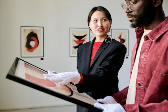 Asian woman and Black man examining artwork in modern gallery setting, wearing professional attire and gloves for protecting exhibits hanging on white walls
