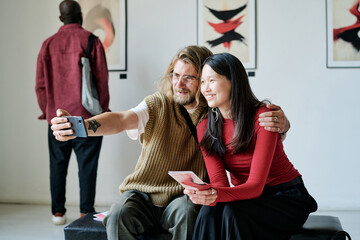 Smiling friends taking selfie in modern art gallery, enjoying artwork in background and capturing their memories sharing memorable moments together