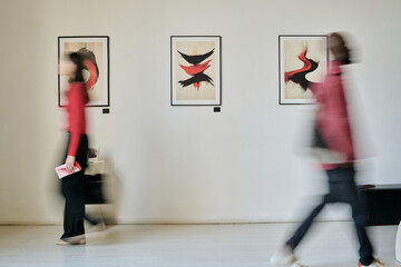 Blurred figures passing by modern art pieces on gallery walls showing dynamic forms and vibrant colors creating sense of motion and interaction with the artwork