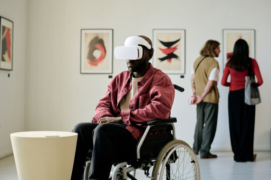 Man in wheelchair experiencing virtual reality headset in modern art gallery, interacting with immersive sculptures on exhibit. Background features diverse visitors engaging with art installations